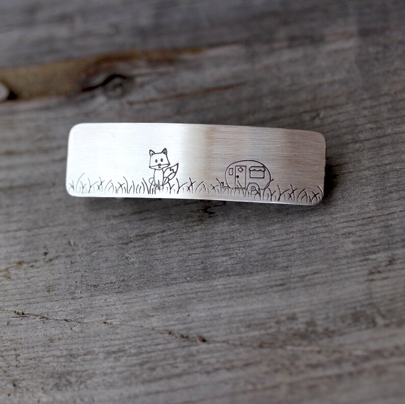 May include: A silver metal hair clip with a fox and a camper engraved on it. The fox is facing the camper and there is grass engraved around them.