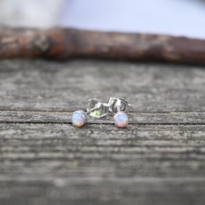 May include: A pair of small stud earrings with iridescent opal-like stones. The earrings are set in silver metal and are displayed on a weathered wooden surface. The opal stones have a soft, pastel color palette.