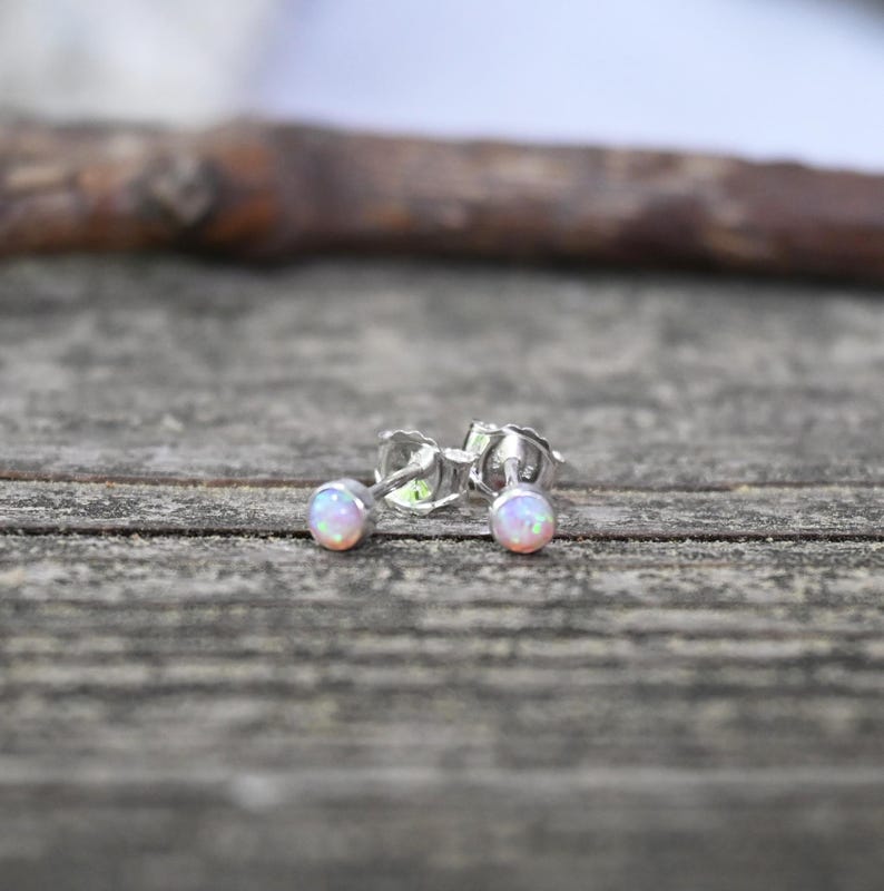 May include: A pair of small stud earrings with iridescent opal-like stones. The earrings are set in silver metal and are displayed on a weathered wooden surface. The opal stones have a soft, pastel color palette.