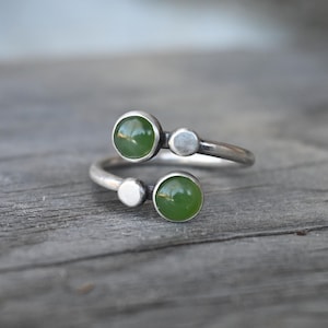 May include: A silver-toned ring featuring two round, vibrant green gemstones and two smaller silver accents. The ring's design is open, with the gemstones positioned on opposite sides of the band. The ring is displayed on a weathered wooden surface.