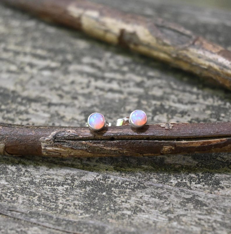May include: A pair of small, round stud earrings with iridescent, opalescent stones. The stones have a soft pink and white color gradient. The earrings are set in silver-colored metal and rest on a wooden branch against a blurred background.