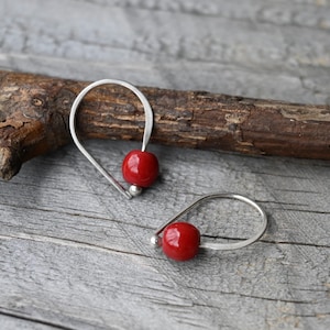 May include: A pair of silver earrings with a teardrop shape. Each earring has a bright red, round bead and a small silver bead. The earrings are presented on a wooden surface.