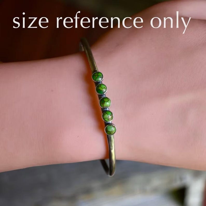 May include: A thin, dark metal cuff bracelet featuring four small, round, green stones with dark veining set in a vertical row along the center. The bracelet has a smooth, oxidized finish and a simple, open-ended design that wraps around the wrist.