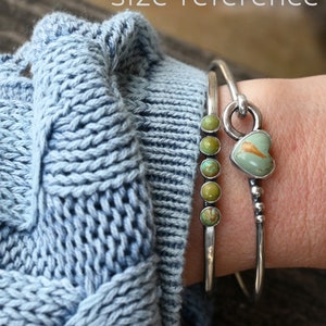 Pode incluir: Duas pulseiras de prata. Uma tem cinco pedras de turquesa verde. A outra tem uma pedra de turquesa em forma de cora&ccedil;&atilde;o. As pulseiras s&atilde;o usadas sobre um su&eacute;ter de malha azul claro. O texto "size reference" est&aacute; no topo.