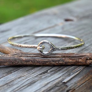 May include: A silver-colored bangle bracelet featuring a hammered, textured surface throughout the band. The bracelet is designed with a circular hook-and-eye style closure mechanism at the center, where one end of the band loops through a ring.