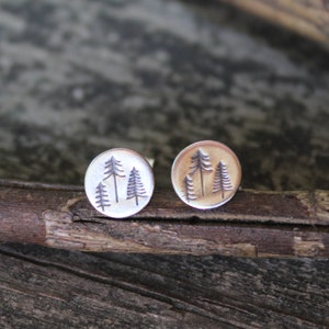 May include: A pair of silver earrings with a design of three pine trees on each earring. The earrings are round and have a slightly textured surface.