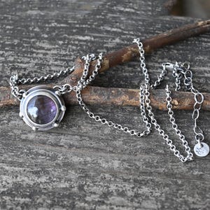 May include: A silver chain necklace with a round pendant featuring a purple gemstone set in a silver bezel. The pendant has a textured silver border.