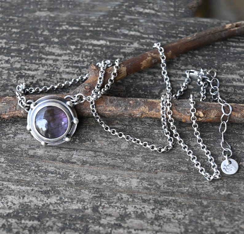 May include: A silver chain necklace with a round pendant featuring a purple gemstone set in a silver bezel. The pendant has a textured silver border.