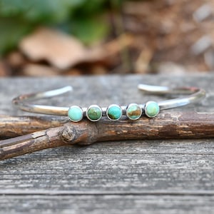 May include: A silver cuff bracelet with five turquoise stones set in a row. The bracelet is on a wooden surface.