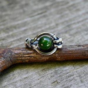 Op de afbeelding: Een zilveren ring met een gepolijste, bolvormige donkergroene edelsteen in een met draad omwikkelde metalen band. De band bestaat uit meerdere strengen zilverdraad die rond de centrale steen buigen en een open, kooiachtige structuur vormen.
