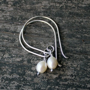 Puede incluir: Un par de pendientes de aro de plata con una sola perla blanca colgando de cada aro.