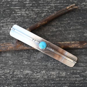 May include: A silver barrette with a turquoise stone in the center. The barrette is rectangular with etched lines and a brushed metal finish. The turquoise stone is round and bright blue.