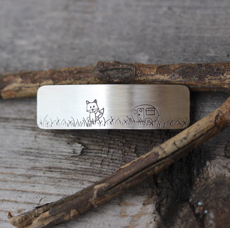 May include: A silver metal bar with an engraved image of a fox and a camper. The fox is standing in a field of grass and the camper is parked behind it.