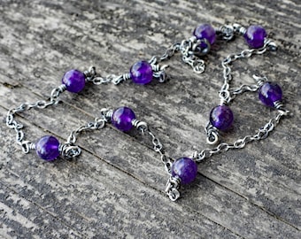 May include: A silver chain necklace with purple amethyst beads. The necklace is on a wooden surface.