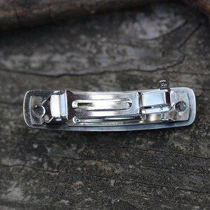 May include: A silver metal hair clip with a smooth, rectangular design. The clip has a spring-loaded mechanism for holding hair in place.