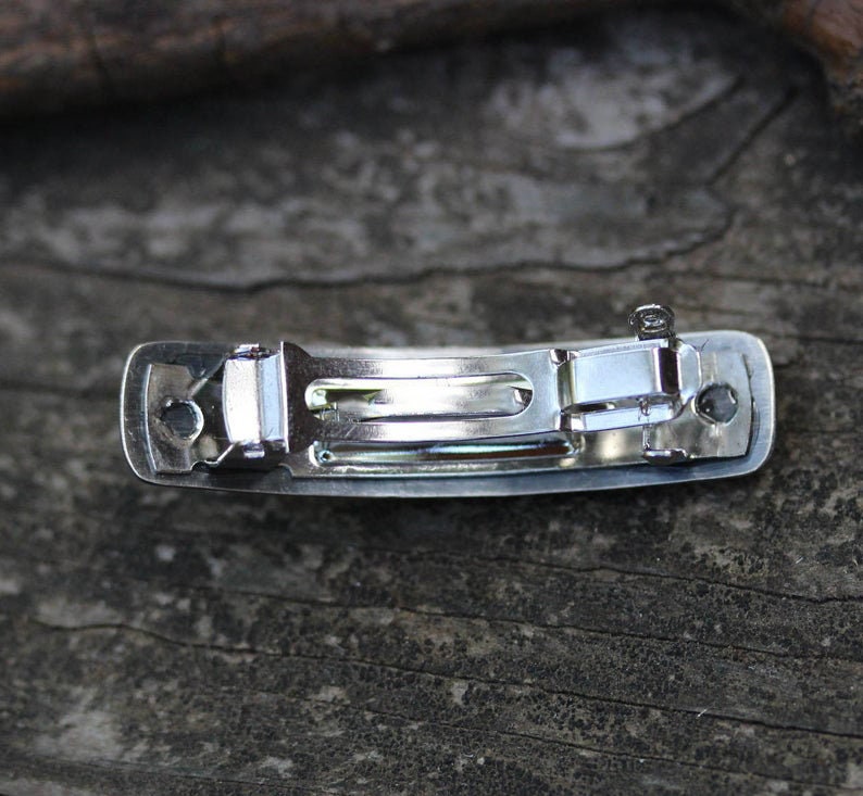 May include: A silver metal hair clip with a smooth, rectangular design. The clip has a spring-loaded mechanism for holding hair in place.