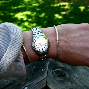 K&ouml;nnte beinhalten: Eine silberne Armbanduhr mit wei&szlig;em Zifferblatt und silbernem Armband. Die Uhr wird an einem Handgelenk getragen, an dem sich auch ein silbernes Armreifarmband befindet.