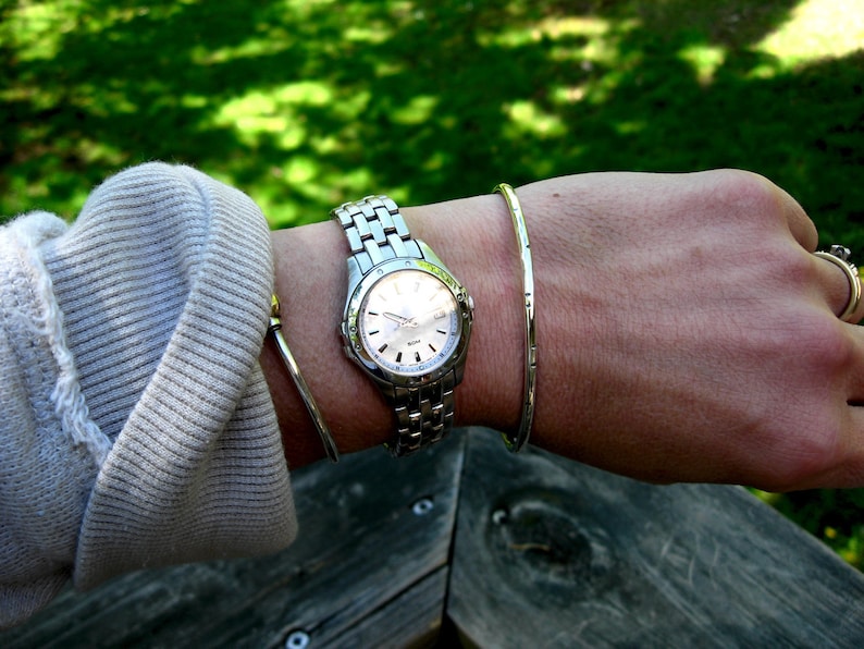 K&ouml;nnte beinhalten: Eine silberne Armbanduhr mit wei&szlig;em Zifferblatt und silbernem Armband. Die Uhr wird an einem Handgelenk getragen, an dem sich auch ein silbernes Armreifarmband befindet.