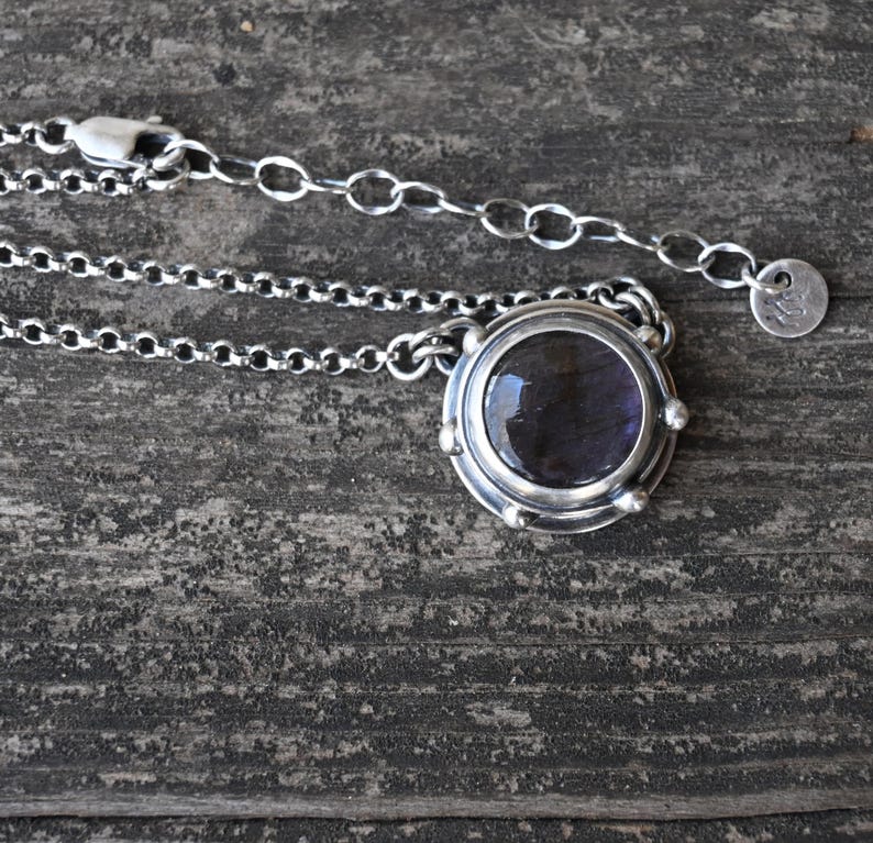 May include: A silver necklace with a round pendant featuring a dark purple gemstone. The pendant has a textured silver border and small silver accents.