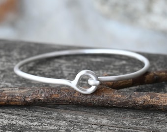 May include: A silver bangle bracelet with a simple, rounded clasp. The bracelet is made of a thin, smooth metal and has a minimalist design.