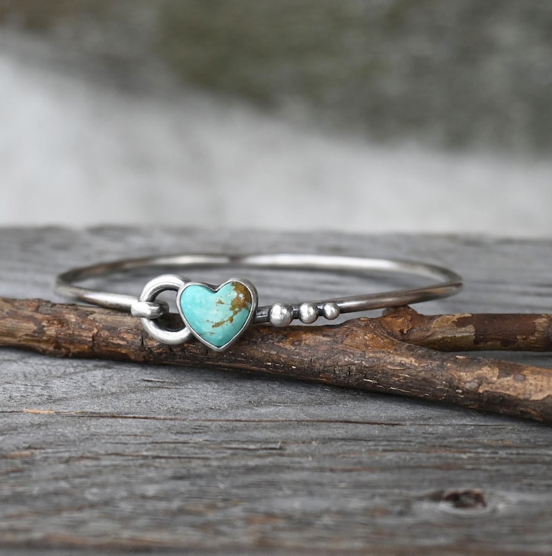 Pode incluir: Uma pulseira de prata com uma pedra turquesa em forma de cora&ccedil;&atilde;o. A pulseira tem um design simples com fecho de gancho e pequenas contas de prata. A pedra turquesa tem veios castanhos.