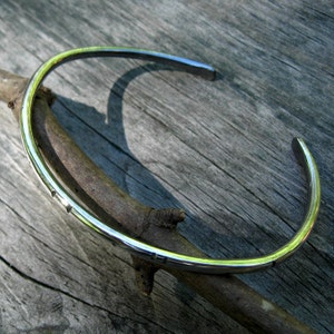 K&ouml;nnte beinhalten: Ein silbernes Armreif-Armband mit glatter, polierter Oberfl&auml;che. Das Armband ist offen und hat ein schlichtes, minimalistisches Design.
