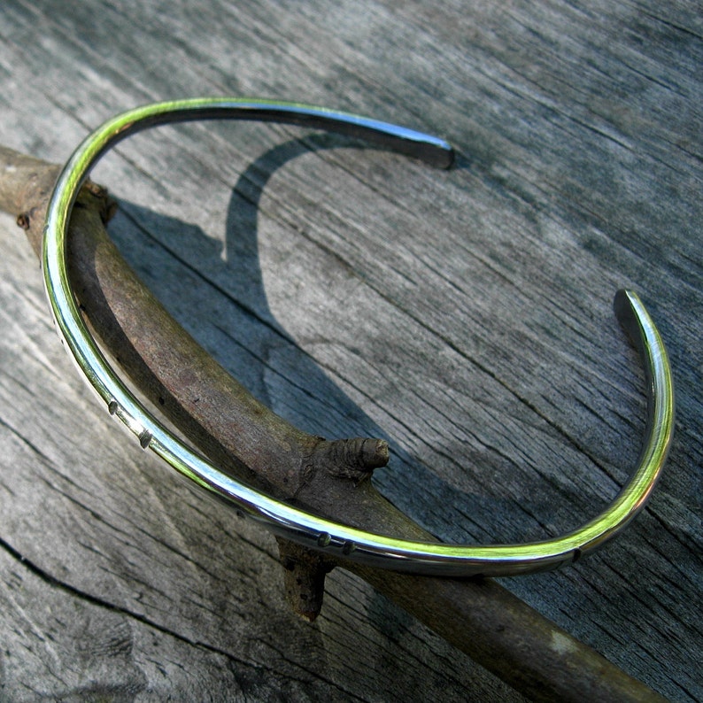 K&ouml;nnte beinhalten: Ein silbernes Armreif-Armband mit glatter, polierter Oberfl&auml;che. Das Armband ist offen und hat ein schlichtes, minimalistisches Design.