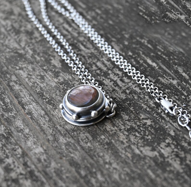 May include: A silver chain necklace with a round pendant featuring a brown and red gemstone set in a silver bezel. The pendant has a textured silver border and three small silver beads.