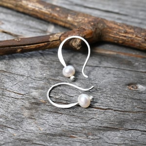 May include: A pair of silver hook earrings, each featuring a single small white pearl and a tiny silver bead at the end of the curved wire. The earrings have a simple, open-loop design that holds the pearl securely in place at the bottom.