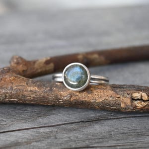 May include: A silver ring with a round, iridescent blue-green gemstone. The ring sits on a piece of wood, showcasing its simple yet elegant design. The gemstone is the focal point, catching the light and displaying a range of colors.