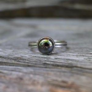May include: A silver-toned ring featuring a smooth, polished, dark gray spherical stone set in a circular bezel. The band of the ring is thin and metallic, showcasing a reflective surface that captures light and surrounding colors in its center.