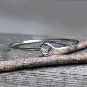 May include: A silver bangle bracelet with a unique, simple, and modern design. The bracelet is made of a thin, smooth metal and features a single, rounded loop that acts as a clasp.