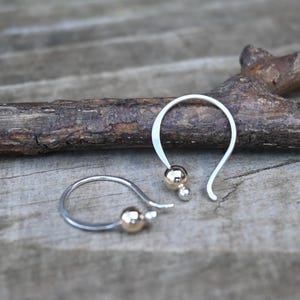 Puede incluir: Un par de pendientes de gancho plateados con detalles de bolas doradas. Los pendientes se exhiben sobre una superficie de madera, mostrando su diseño simple pero elegante. Los pendientes tienen un diseño de gancho curvo con una pequeña bola dorada y una bola dorada ligeramente más pequeña.