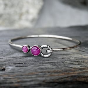 Pode incluir: Uma pulseira de prata com duas pedras preciosas redondas rosa brilhante e um fecho circular. A pulseira é exibida sobre uma superfície de madeira rústica, destacando o contraste entre o metal polido e a textura natural.