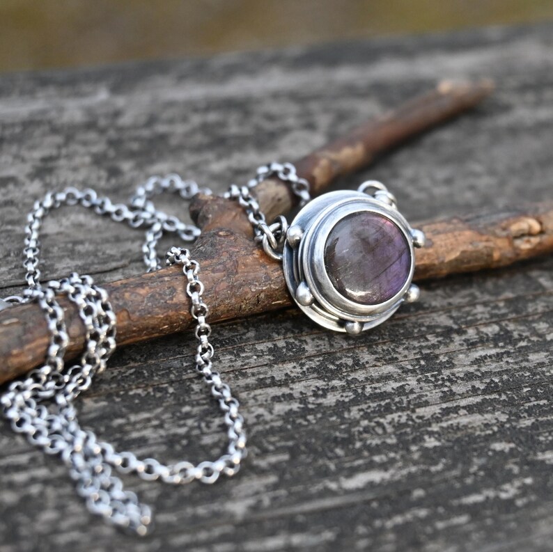 May include: A silver necklace with a round pendant featuring a purple gemstone set in a silver bezel. The pendant has a textured silver border and small silver beads around the edge.