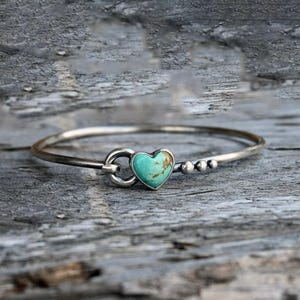 Pode incluir: Uma pulseira de bracelete prateada com uma pedra turquesa em forma de cora&ccedil;&atilde;o. A pulseira tem um fecho de gancho e pequenas contas prateadas. A pedra turquesa tem veios castanhos.