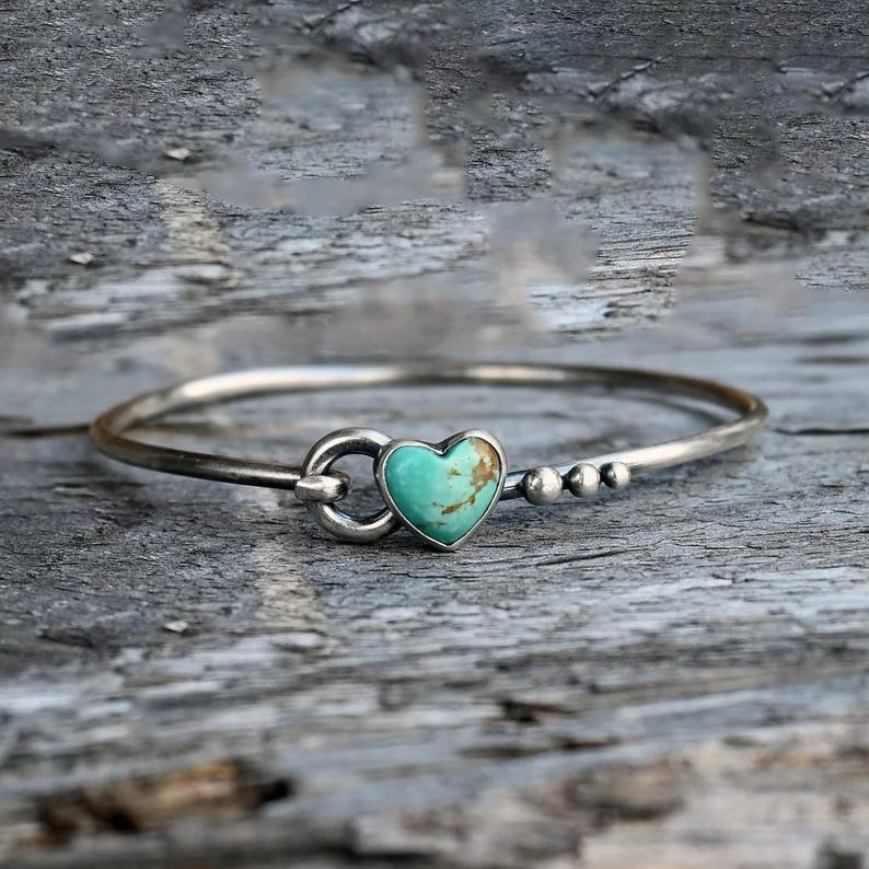Pode incluir: Uma pulseira de bracelete prateada com uma pedra turquesa em forma de cora&ccedil;&atilde;o. A pulseira tem um fecho de gancho e pequenas contas prateadas. A pedra turquesa tem veios castanhos.
