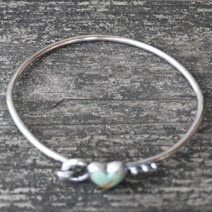 Pode incluir: Uma pulseira de prata com um berloque em forma de cora&ccedil;&atilde;o. O cora&ccedil;&atilde;o &eacute; verde claro com detalhes em castanho. A pulseira tem um design de n&oacute; de um lado e pequenas contas de prata do outro. A pulseira est&aacute; sobre uma superf&iacute;cie de madeira cinza.