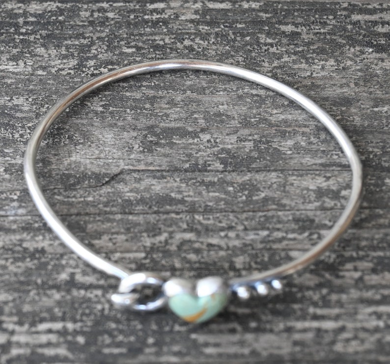 Pode incluir: Uma pulseira de prata com um berloque em forma de cora&ccedil;&atilde;o. O cora&ccedil;&atilde;o &eacute; verde claro com detalhes em castanho. A pulseira tem um design de n&oacute; de um lado e pequenas contas de prata do outro. A pulseira est&aacute; sobre uma superf&iacute;cie de madeira cinza.