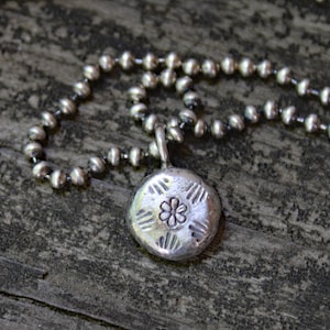 May include: A silver necklace with a round pendant featuring a stylised flower design. The pendant is attached to a silver chain with small round beads.