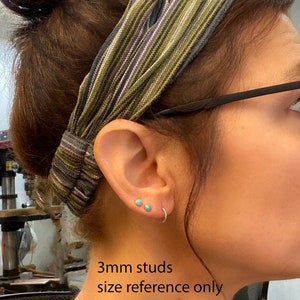 May include: A close-up of a person's ear with two small, turquoise stud earrings and a silver hoop earring. The text "3mm studs size reference only" is visible. The person is wearing a striped headband.