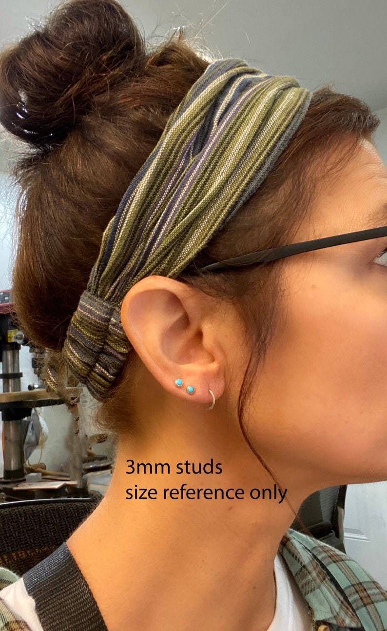 May include: A close-up of a person's ear with two small, turquoise stud earrings and a silver hoop earring. The text "3mm studs size reference only" is visible. The person is wearing a striped headband.