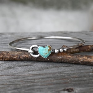 Pode incluir: Uma pulseira de prata com uma pedra turquesa em forma de cora&ccedil;&atilde;o. A pulseira tem um design simples e elegante com um fecho e pequenas contas de prata. A pedra turquesa tem inclus&otilde;es castanhas. A pulseira &eacute; exibida numa superf&iacute;cie de madeira.