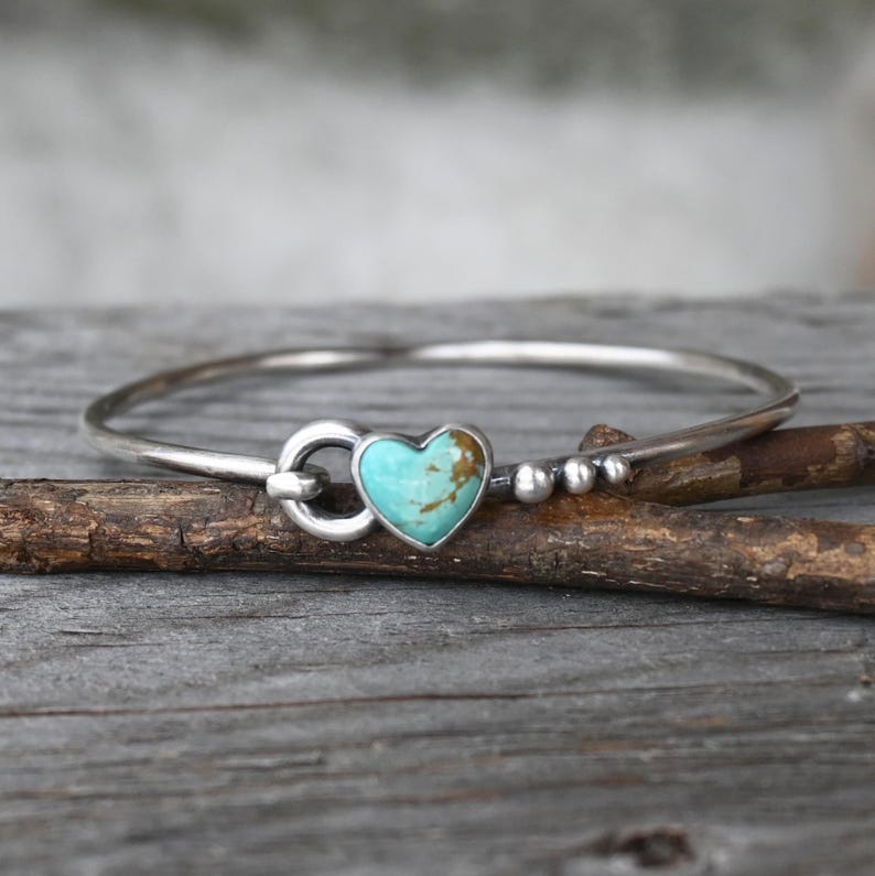 Pode incluir: Uma pulseira de prata com uma pedra turquesa em forma de cora&ccedil;&atilde;o. A pulseira tem um design simples e elegante com um fecho e pequenas contas de prata. A pedra turquesa tem inclus&otilde;es castanhas. A pulseira &eacute; exibida numa superf&iacute;cie de madeira.