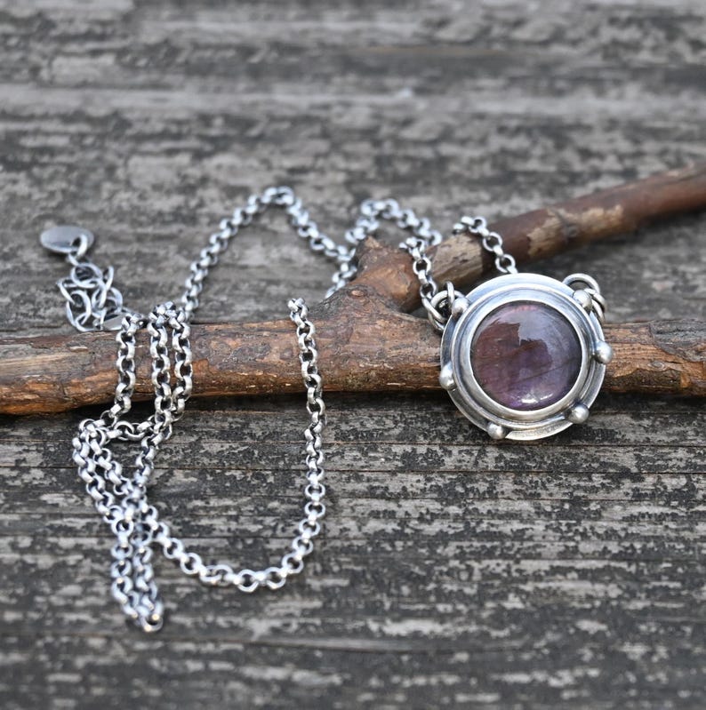 May include: A silver necklace with a round pendant featuring a purple gemstone set in a silver bezel. The pendant has a decorative border and is attached to a silver chain.