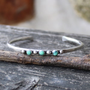 May include: A silver bangle bracelet with three turquoise stones set in a row. The bracelet is on a wooden surface.