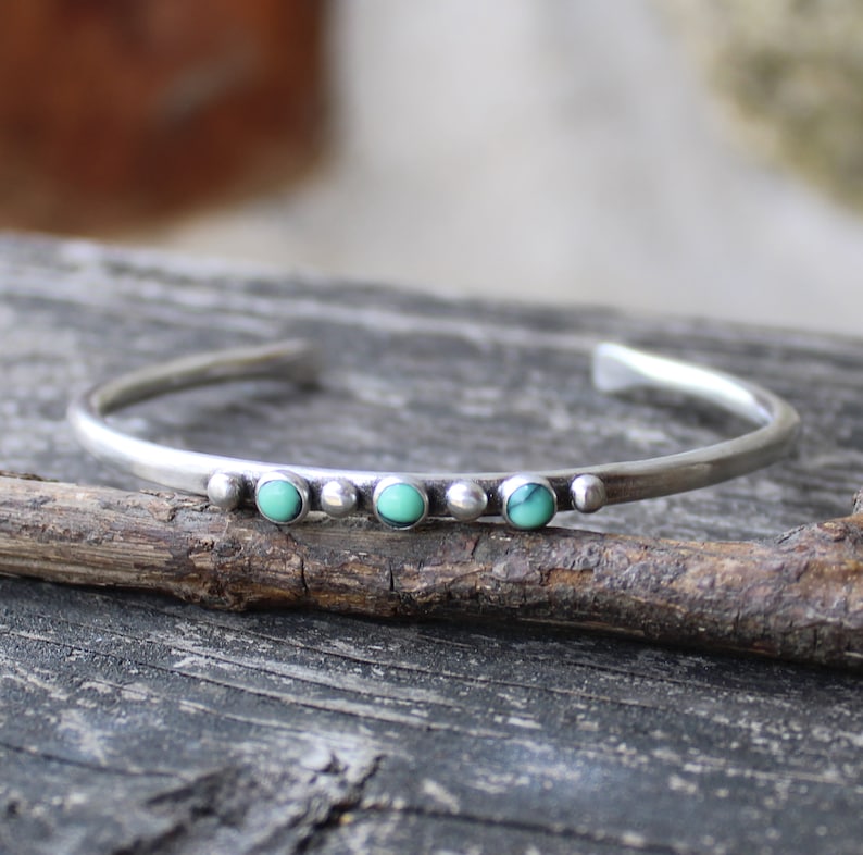 May include: A silver bangle bracelet with three turquoise stones set in a row. The bracelet is on a wooden surface.