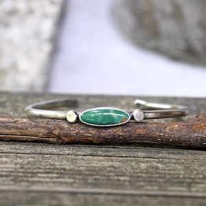 May include: A silver cuff bracelet featuring a vibrant green oval turquoise stone at the center. The bracelet has a minimalist design with small, round accents on either side of the turquoise. The bracelet is displayed on a wooden surface.