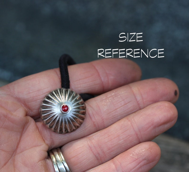 May include: A silver hair tie with a red gemstone in the center. The hair tie is in the shape of a dome with a ribbed design.