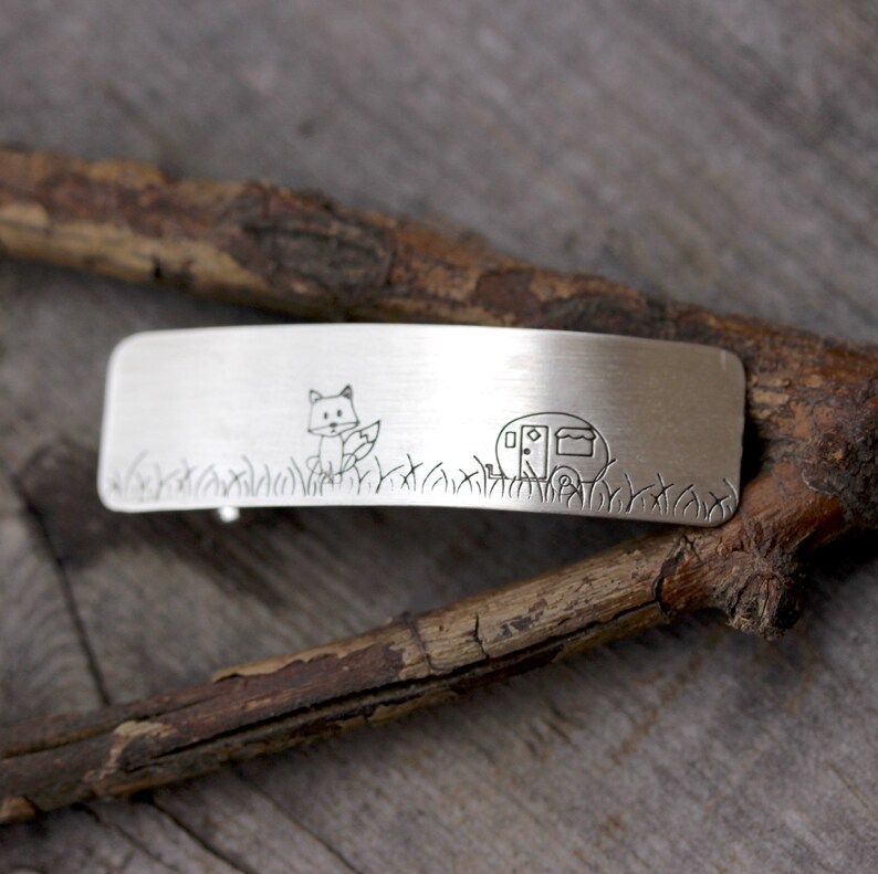 May include: Silver hair clip with a fox and a camper engraved on it. The clip is on a wooden surface.